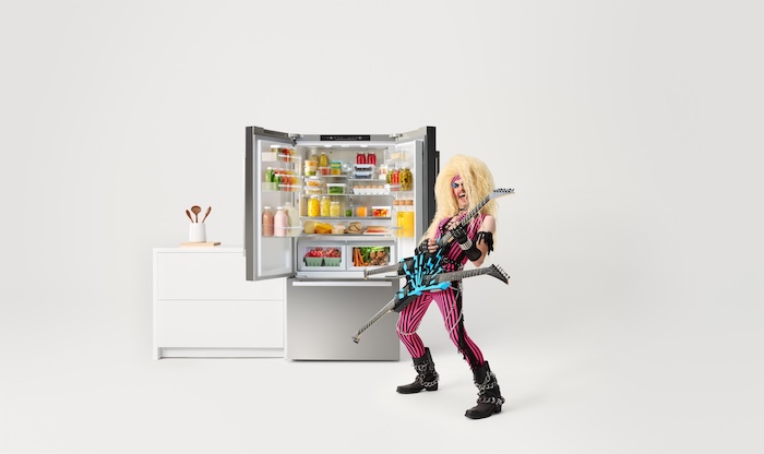 A Bosch 800 Series French-Door Refrigerator is highlighted alongside a classic 70s rockstar. The rocker, dressed in colorful clothes with wild blonde hair, passionately plays an electric guitar, creating an edgy and innovative atmosphere