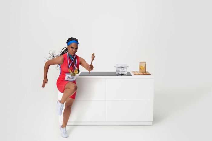 A dynamic woman in a sprinter pose and athletic wear poses on a modern 800 series induction cooktop, showcasing its sleek design and powerful performance